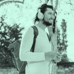 Jovem homem com mochila nas costas e fones de ouvidos em parque desfocado. Supõ-se que está ouvindo áudio livros e podcasts