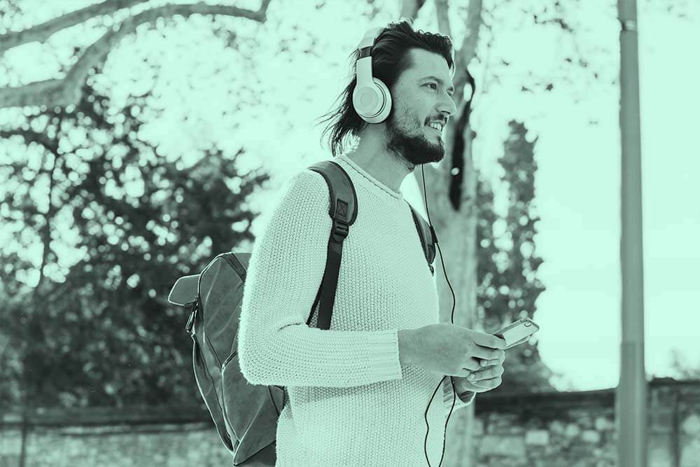 Jovem homem com mochila nas costas e fones de ouvidos em parque desfocado. Supõ-se que está ouvindo áudio livros e podcasts