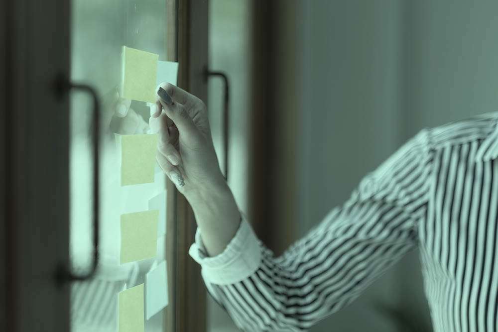 Close em braço e mão feminina fixando postit em vidro para check list de afiliados