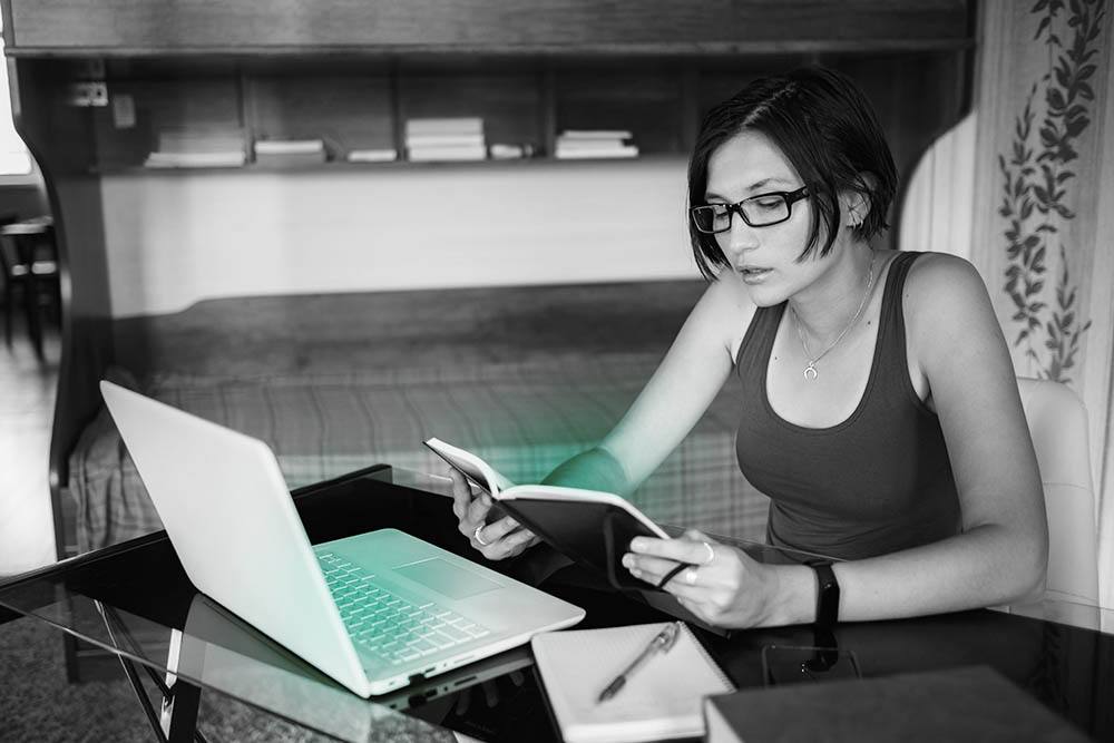 Jovem mulher lendo um caderno que segura diante de tela de laptop que projetam luz verde