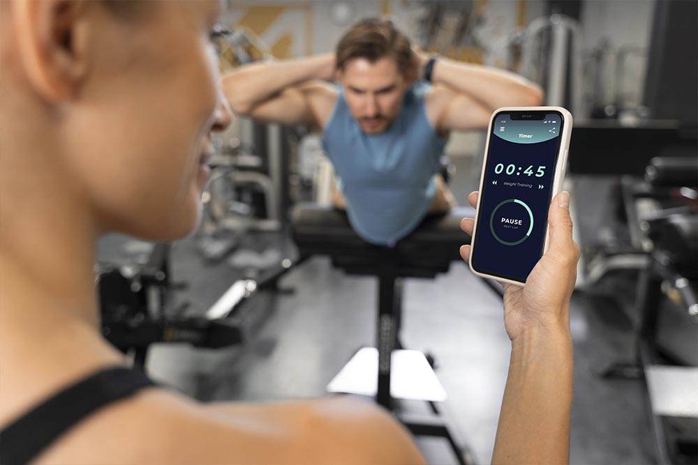 Close no celular com aplicativo fitness nas mãos de jovem mulher e ao fundo rapaz fazendo abdominais em ambiente de academia