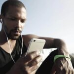 Homem com blusa regata e calça esportiva sentado no chão ao ar livre com fones de ouvido olhando a tela do celular