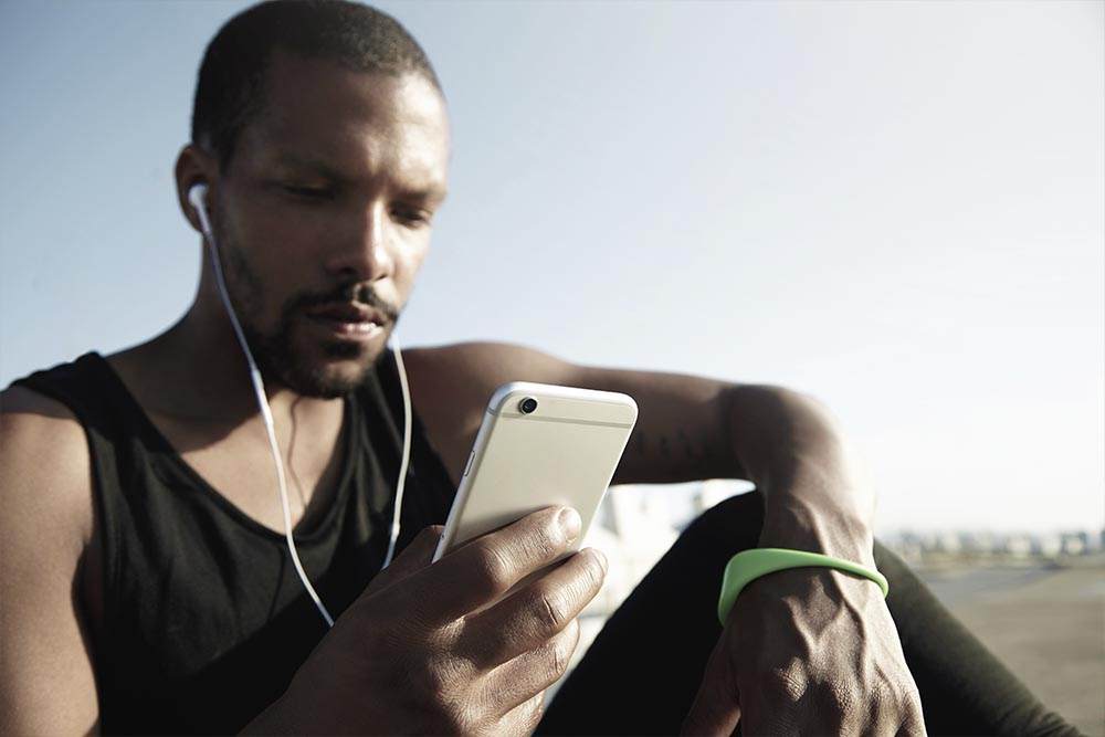 Homem com blusa regata e calça esportiva sentado no chão ao ar livre com fones de ouvido olhando a tela do celular