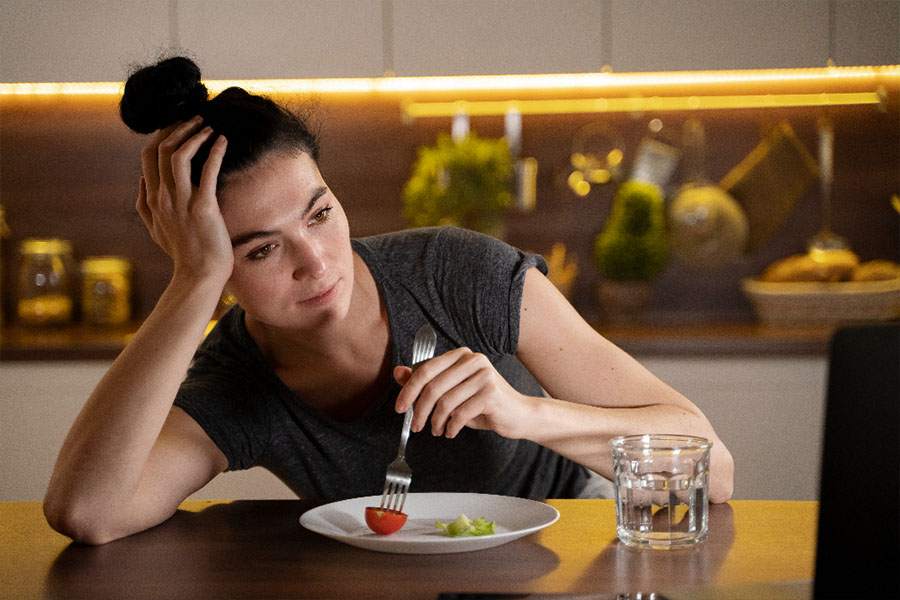 Mulher atrás de uma mesa diante de um prato com pequena salada segurando o garfo com uma mão e a cabeça apoiada na outra mão. A expressão de desânimo representa o sintoma de TARE