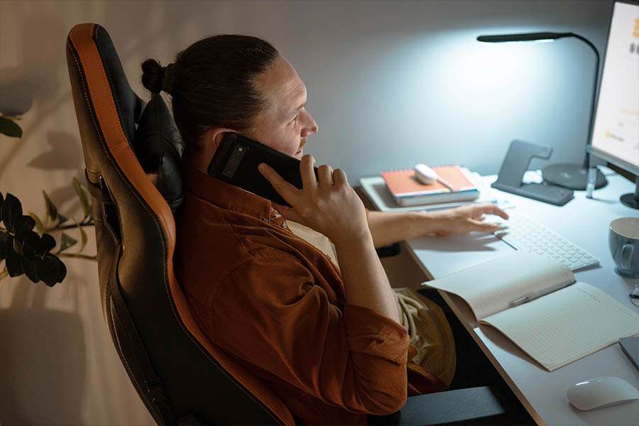 Homem recostado em cadeira gamer com um braço sobre a mesa de escritório e o outro segura um celular junto ao ouvido. Uma luminária compõe um ambiente confortável ergonômico.