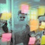 Vemos por uma janela de vidro três Jovens sorridentes observando Estratégias de Marketing em Post-it fixados