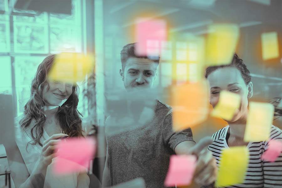 Vemos por uma janela de vidro três Jovens sorridentes observando Estratégias de Marketing em Post-it fixados